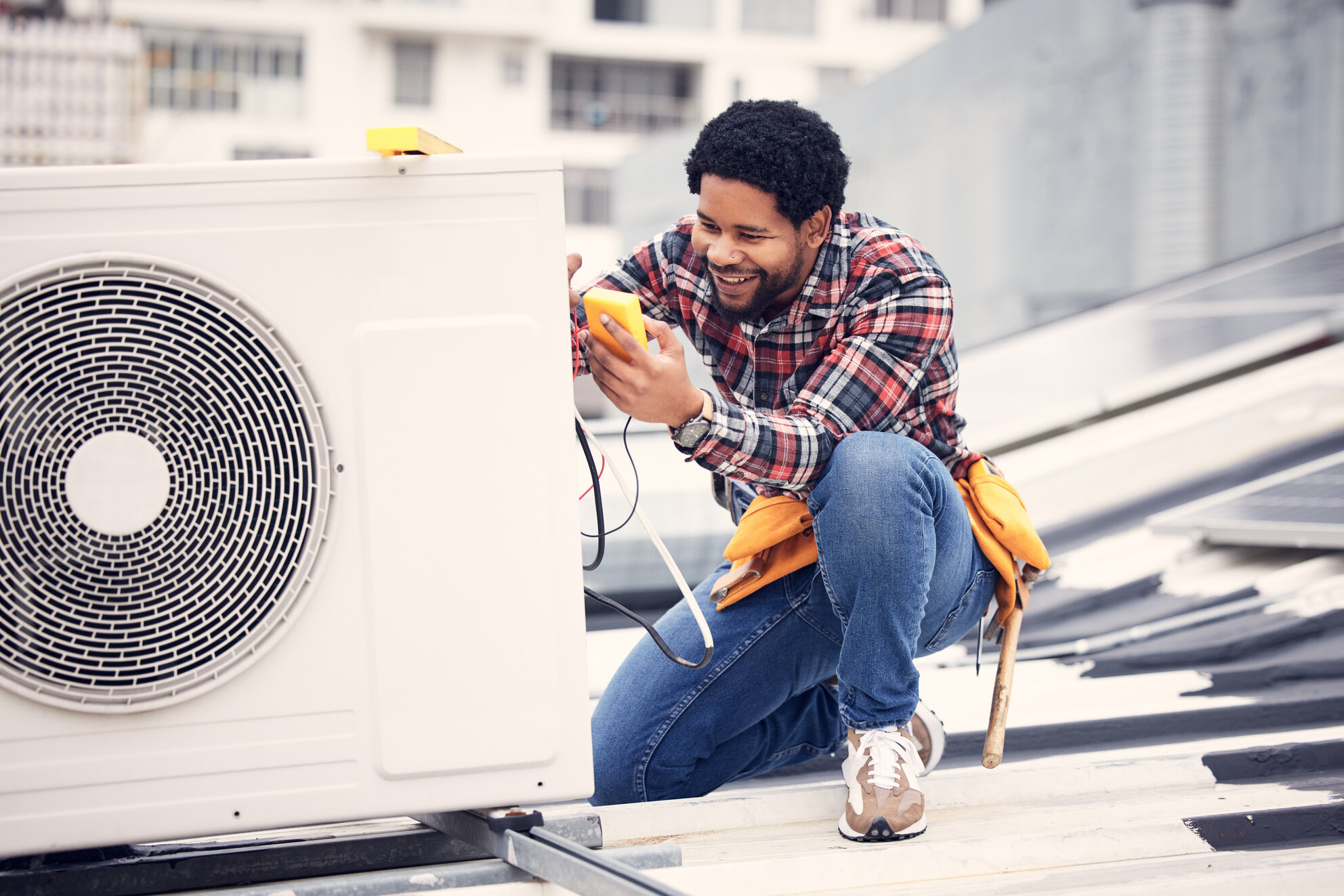 Technician performing maintenance on an air conditioning unit, using a multimeter to check electrical components, on a rooftop setting, emphasizing HVAC service and repair expertise.